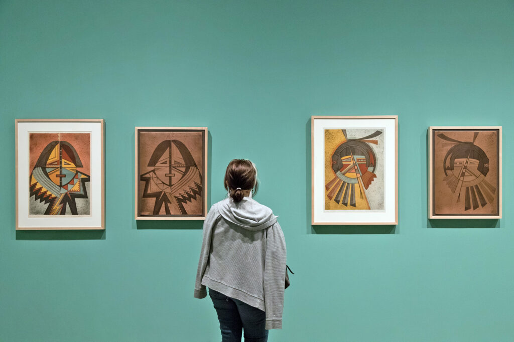 A person stands in front of four framed abstract portraits displayed on a green wall, reflecting on the gallery’s past exhibitions.