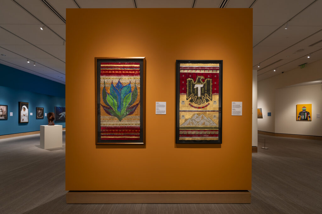 Two framed textile artworks hang on an orange gallery wall, each featuring colorful patterns and icons and symbols, with exhibition labels beside them in a modern museum setting.