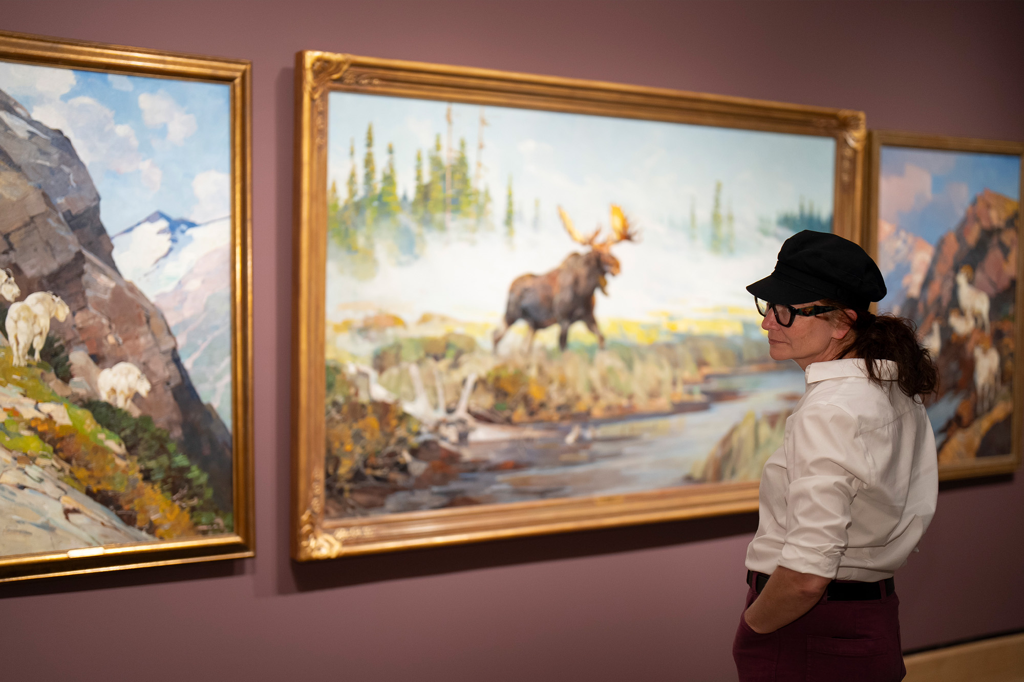 A person wearing a white shirt, dark pants, and a black cap views a large painting of moose wildlife in a natural landscape at an art gallery.
