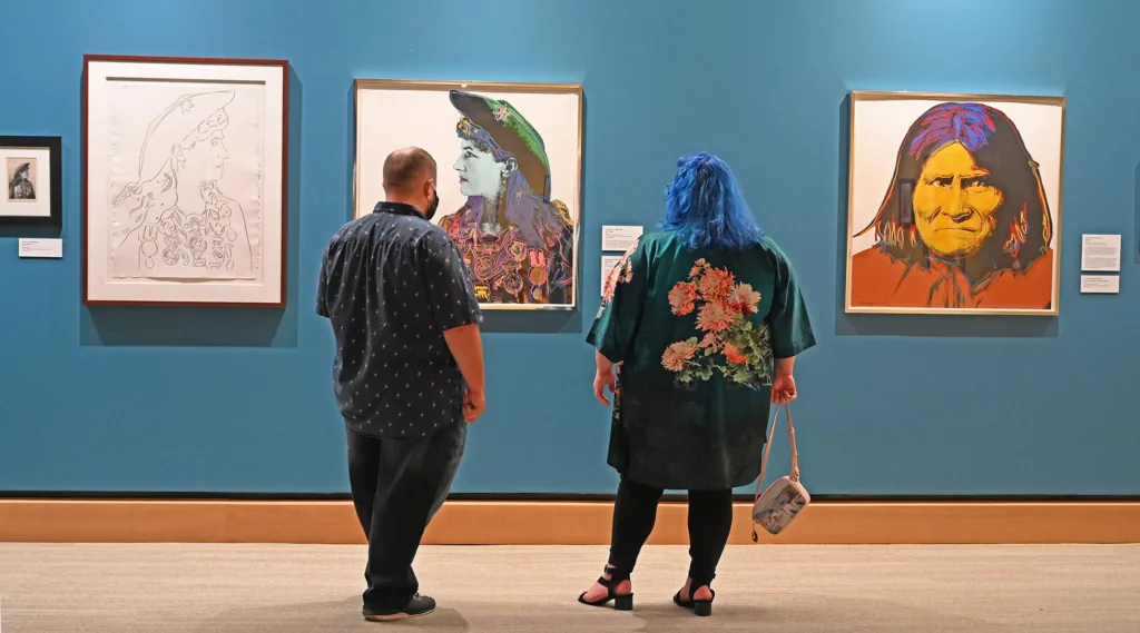 Two people stand in front of three colorful portraits displayed on a blue wall in an art gallery, reminiscent of Warhol's West and the vibrant legacy of Western art.