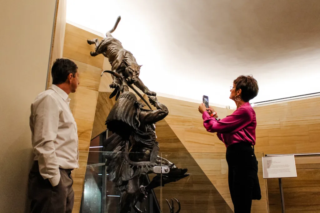 A woman photographs a large sculpture of a man being attacked by a wildcat, while a man looks on in a museum setting, capturing the dramatic scene amidst exhibits showcasing moments from celebrations and weddings.
