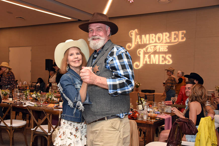 An older couple in western attire dances at an indoor event, with a "Jamboree at The James" sign projected on the wall behind them.