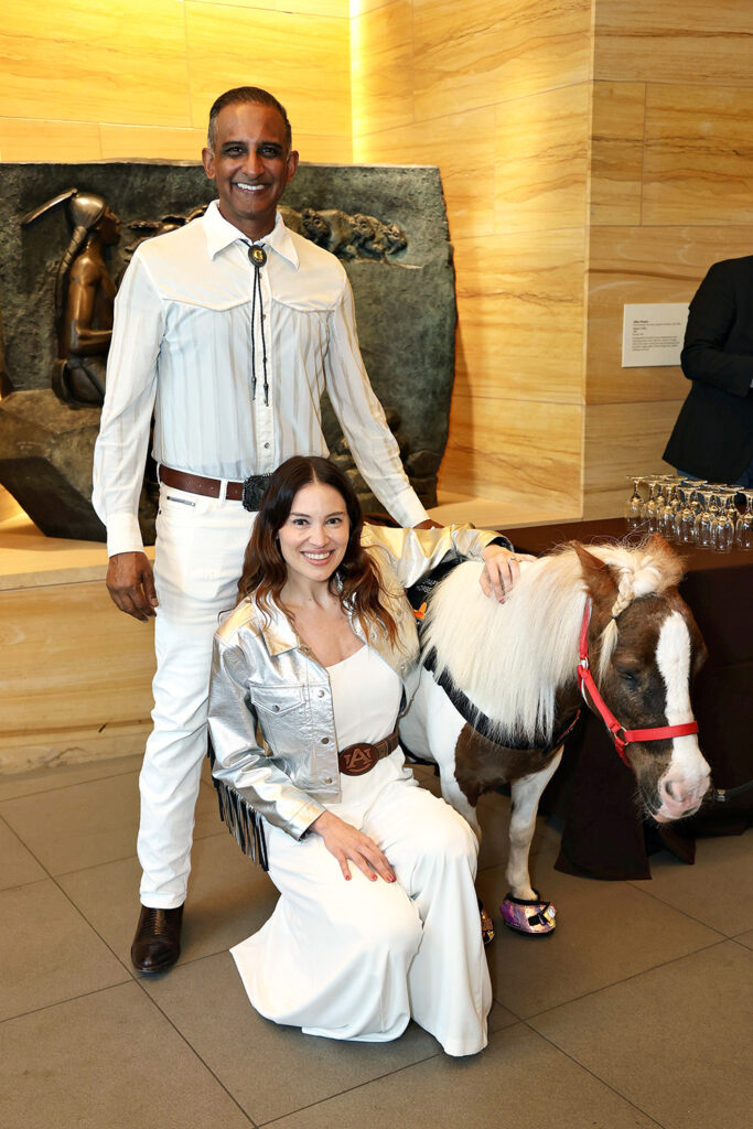 A man and a woman in white outfits pose indoors with a small pony wearing a red halter and decorative shoes at The James jamboree. A sculpture and refreshments are visible in the background.