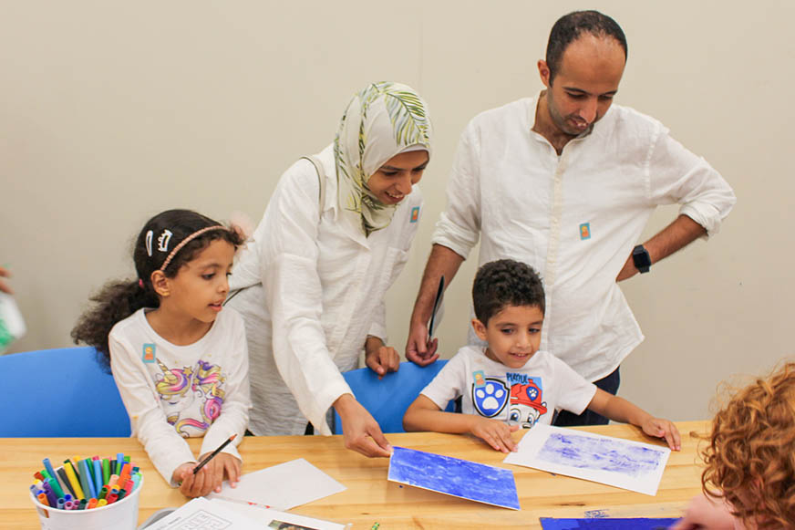 A family of four, including two adults and two children, participate in family programs as they work on art projects at a table with markers, papers, and drawings displayed.