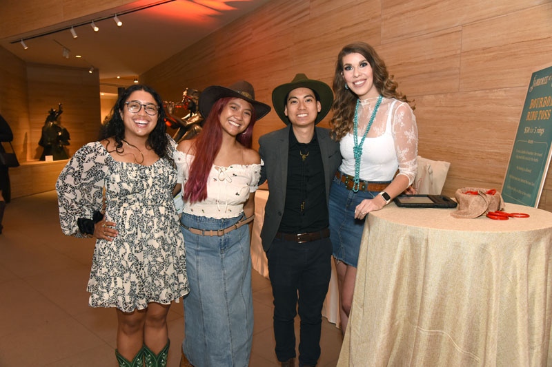 Four people dressed in western-themed attire stand together and smile by a round table at The James during the lively Jamboree, all set in a modern indoor setting.