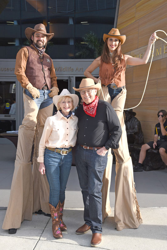 Two people on stilts dressed as cowboys stand behind an older couple in Western attire, posing together outside The James during the lively Jamboree.