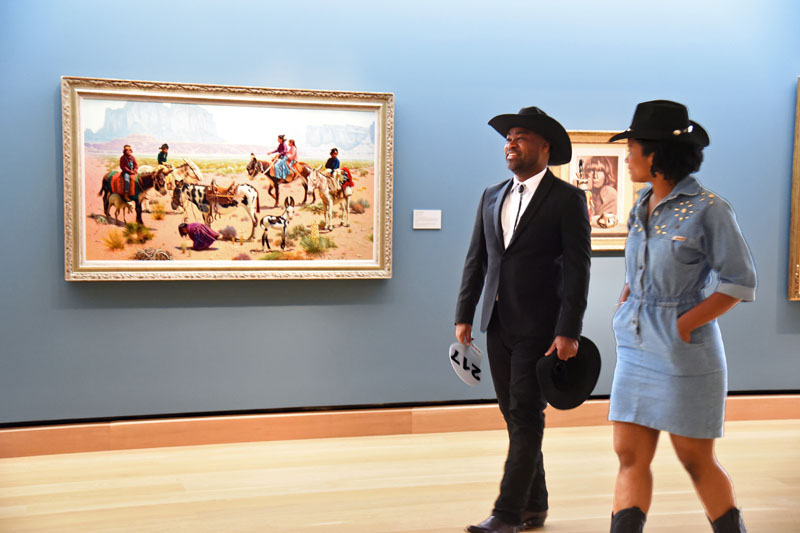 Two people in Western attire walk through The James art gallery, passing a large painting of cowboys and horses in a desert landscape, evoking the spirit of a timeless jamboree.