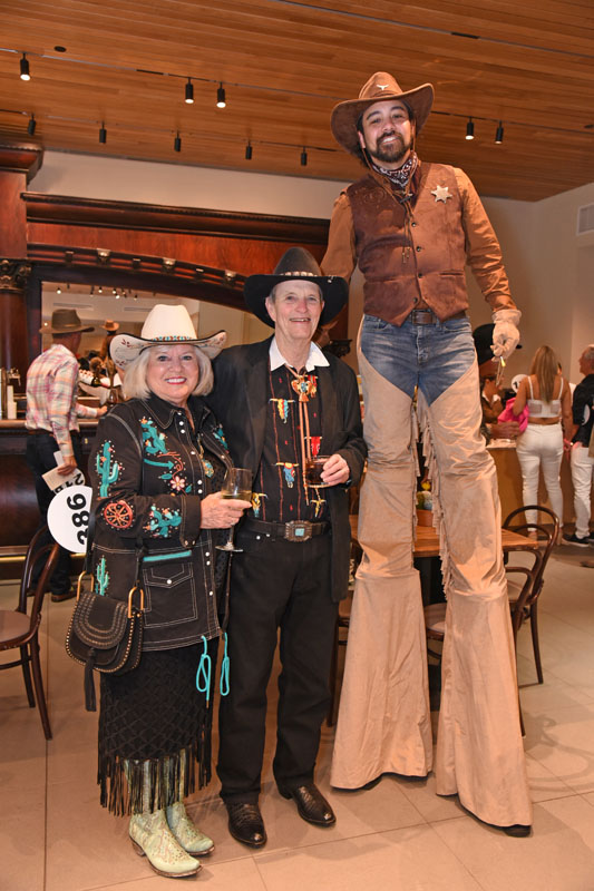 Three adults in Western attire pose indoors at The James; one man is on stilts, while the other two stand side by side holding drinks and smiling, giving a festive jamboree vibe.