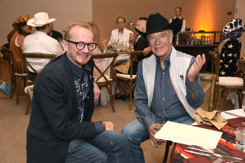 Two men sit and smile at a table during The James Jamboree; one wears a black cowboy hat and the other glasses and a blazer. Other guests mingle in the background.