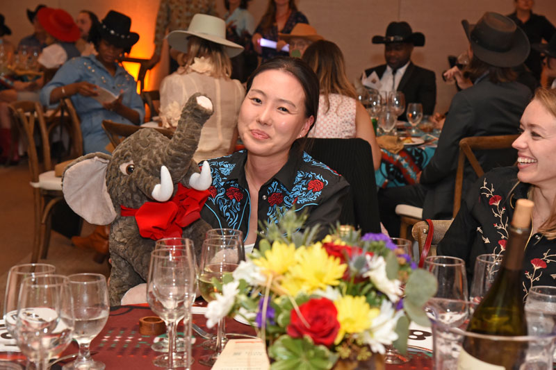 At The James Jamboree, a woman in western attire sits at a table with empty wine glasses, holding a plush elephant toy, surrounded by people in cowboy hats at the lively themed event.