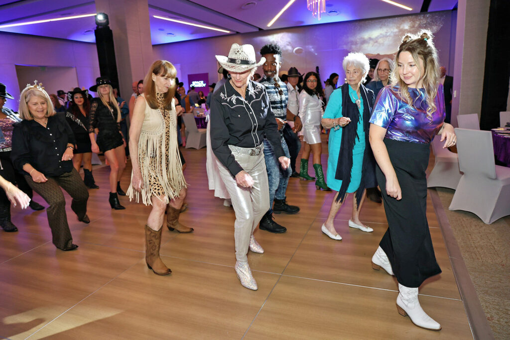 A group of people dressed in themed outfits dance in formation at The James Jamboree, held in a decorated indoor venue with tables and chairs in the background.