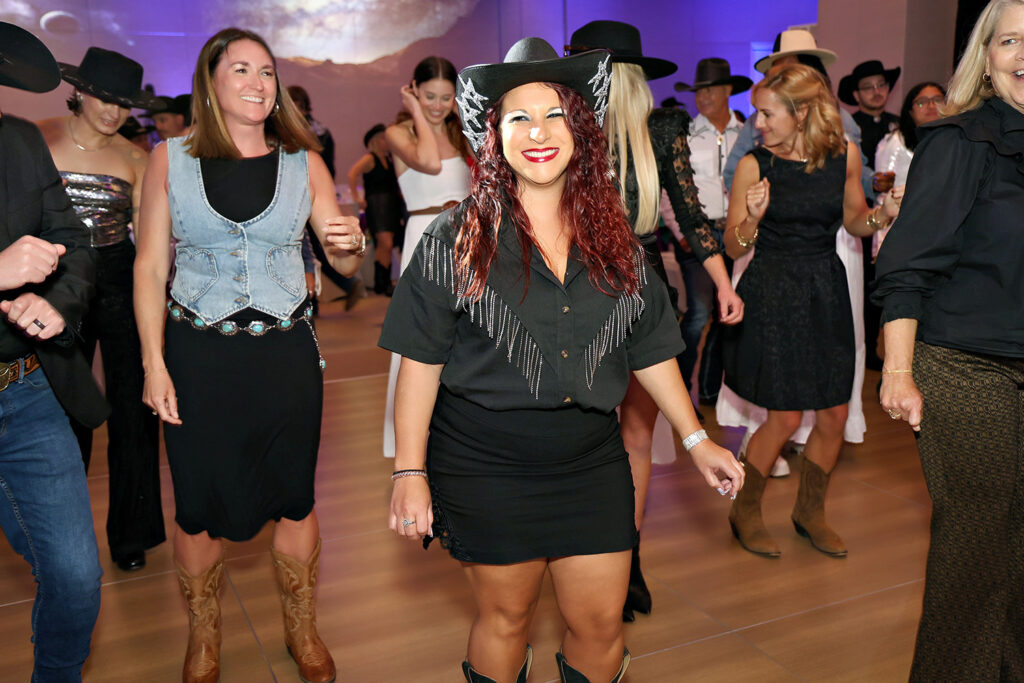 People in western-style outfits, including cowboy hats and boots, dance together indoors under bright lighting at The James Jamboree.
