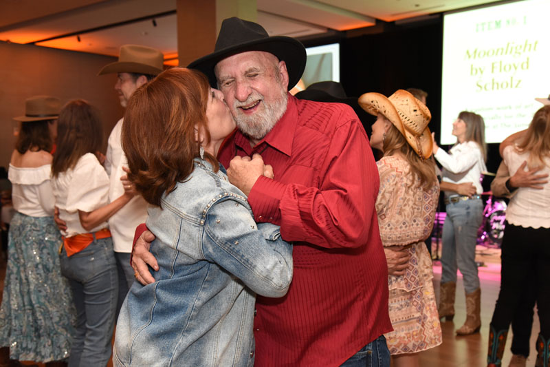 A woman kisses a smiling man in a red shirt and black hat while dancing at The James Jamboree, a lively western-themed event with others in cowboy attire enjoying the festivities in the background.