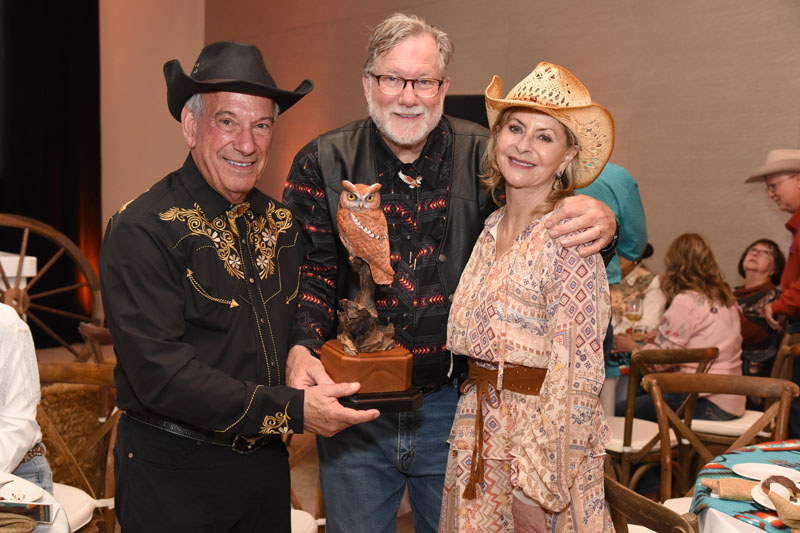 Three people in Western attire pose indoors at The James Jamboree, with one man holding an owl-shaped trophy. Tables and guests fill the background.