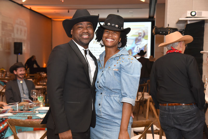 Two people in cowboy hats smile and pose for a photo at an indoor Jamboree event. Other attendees are visible in the background.