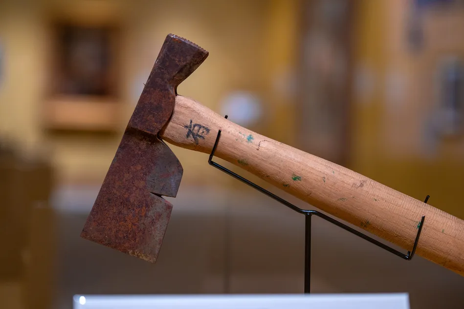 A close-up of a rusty metal axe head with a distinctive notch, attached to a wooden handle, displayed on a metal stand in a museum setting, highlights tools from Far East 02 Mining.