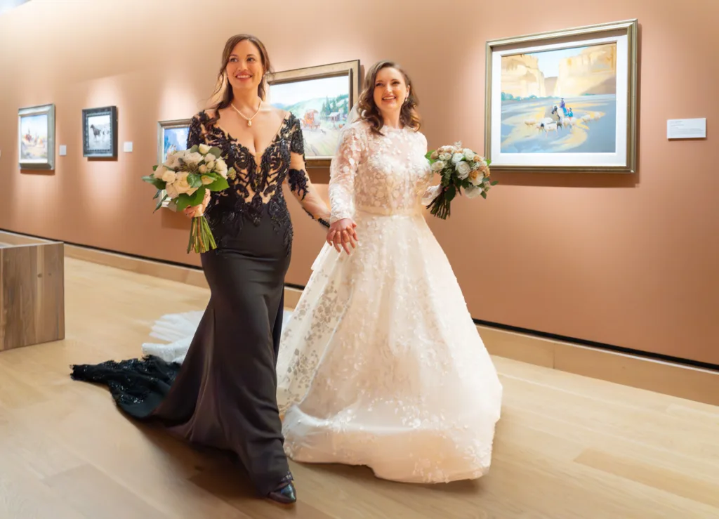 Two brides, one in a black gown and one in a white gown, hold hands and carry bouquets while walking through an art gallery, capturing the elegance of weddings and the joy of celebrations.
