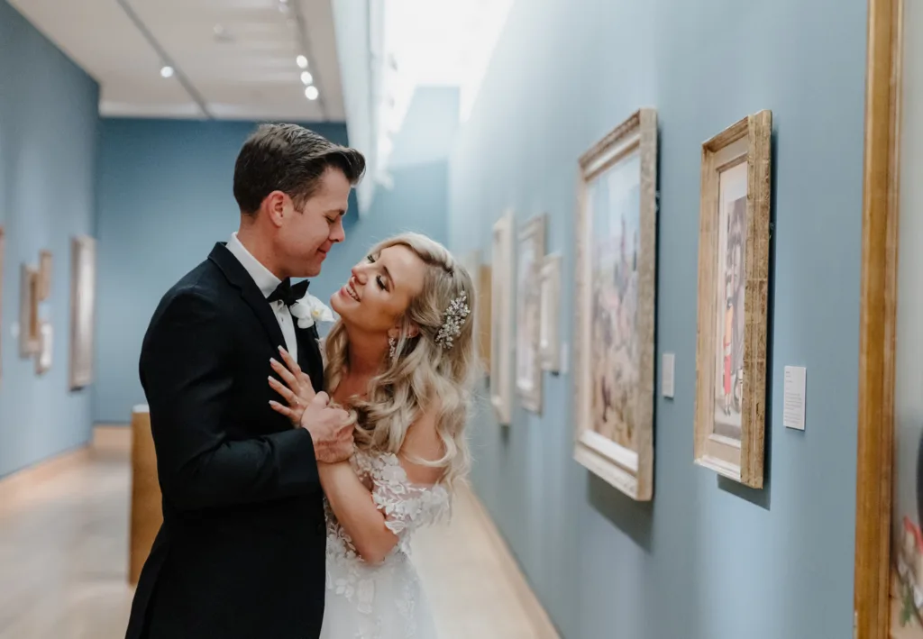 A bride and groom in wedding attire smile and embrace inside an art gallery with framed paintings on blue walls, capturing the joy of weddings and celebrations.