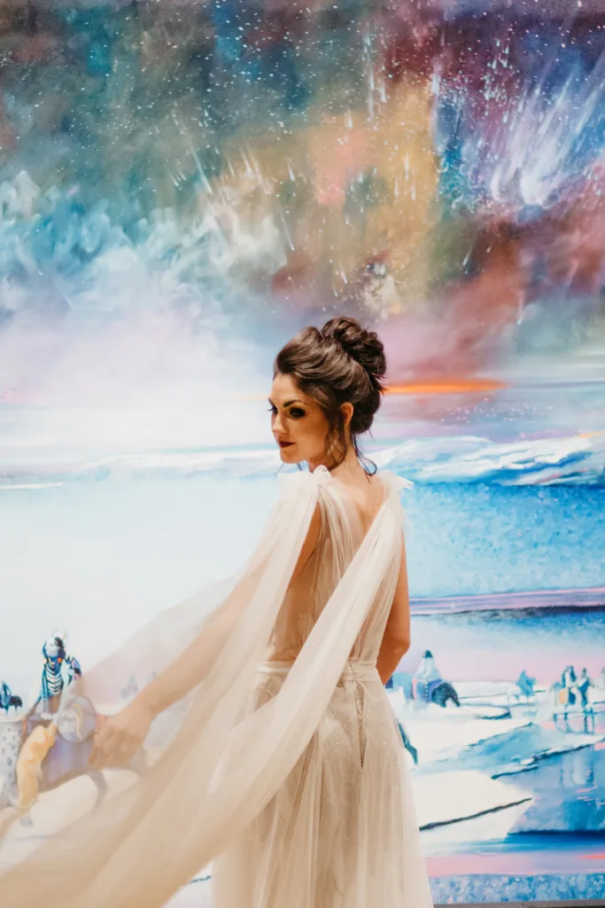 A woman in a flowing white gown stands before a colorful, abstract painting with cosmic and icy landscape elements, capturing the enchanting spirit of weddings and celebrations.
