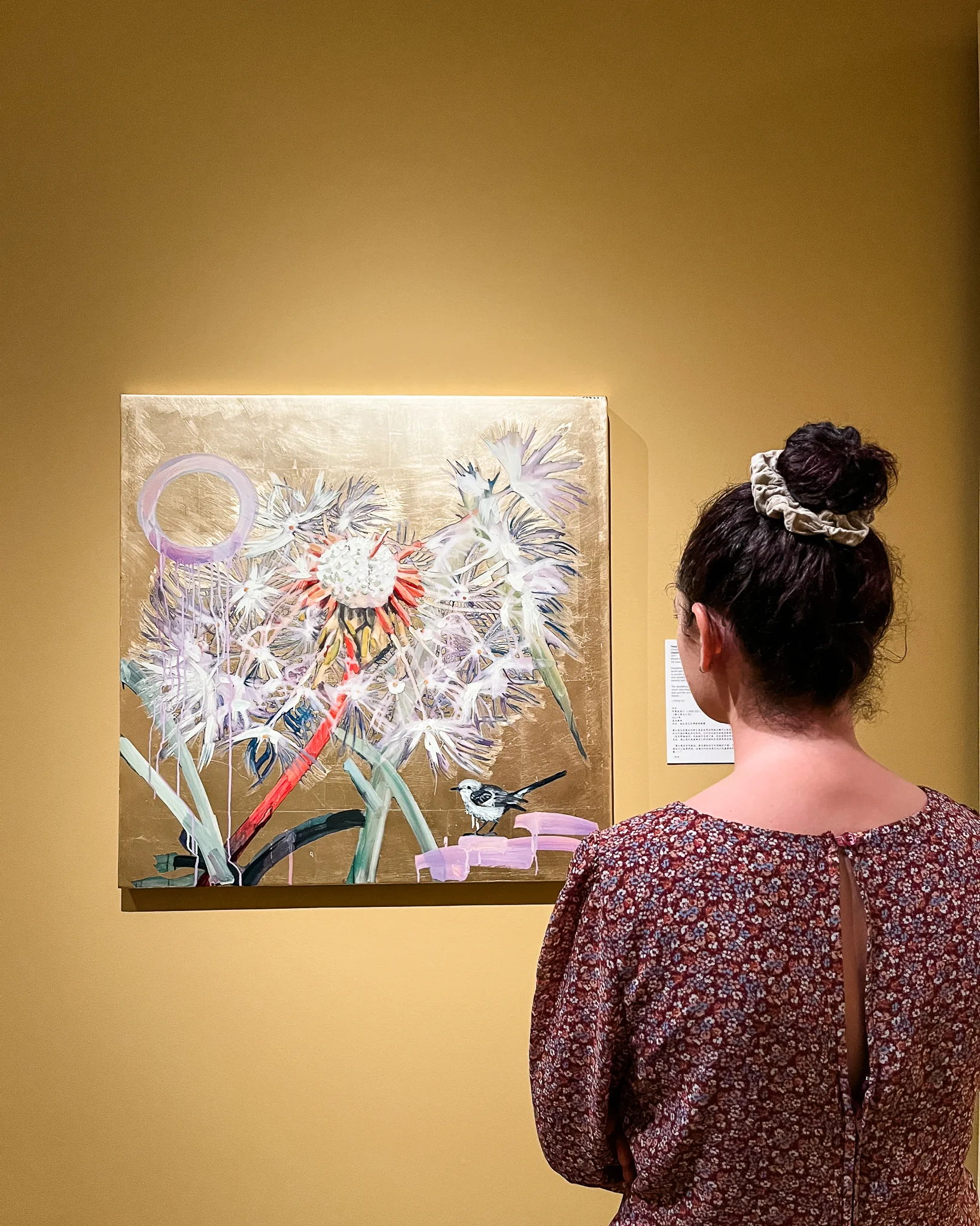 A woman with her hair in a bun stands in front of the FarEast 12 Reflections floral painting on a yellow wall, admiring it in an art gallery.