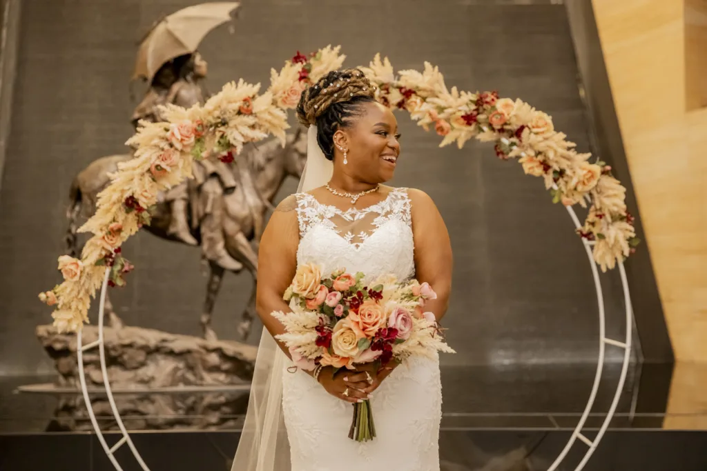 A bride in a white lace gown holds a bouquet and stands in front of a floral arch, with a horse rider statue in the background—a timeless moment perfect for weddings and celebrations.