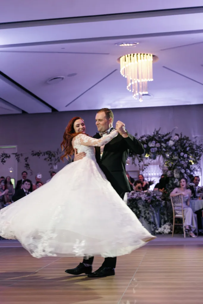 A bride and groom dance together at their wedding reception, with guests seated and watching in the background and floral decorations behind them, capturing the joy of weddings and celebrations.