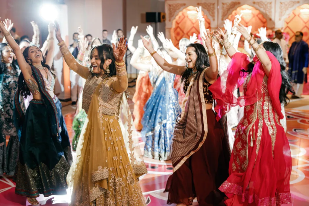 A group of women in colorful traditional Indian attire dance together with raised arms at an indoor wedding celebration.