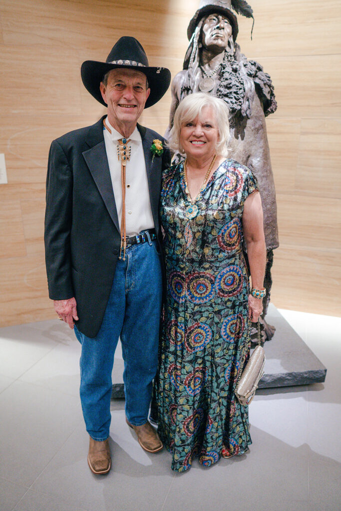 An older man in a cowboy hat and bolo tie stands beside a woman in a colorful dress, both smiling in front of a Native American statue, highlighting the founders' enduring role in company history.