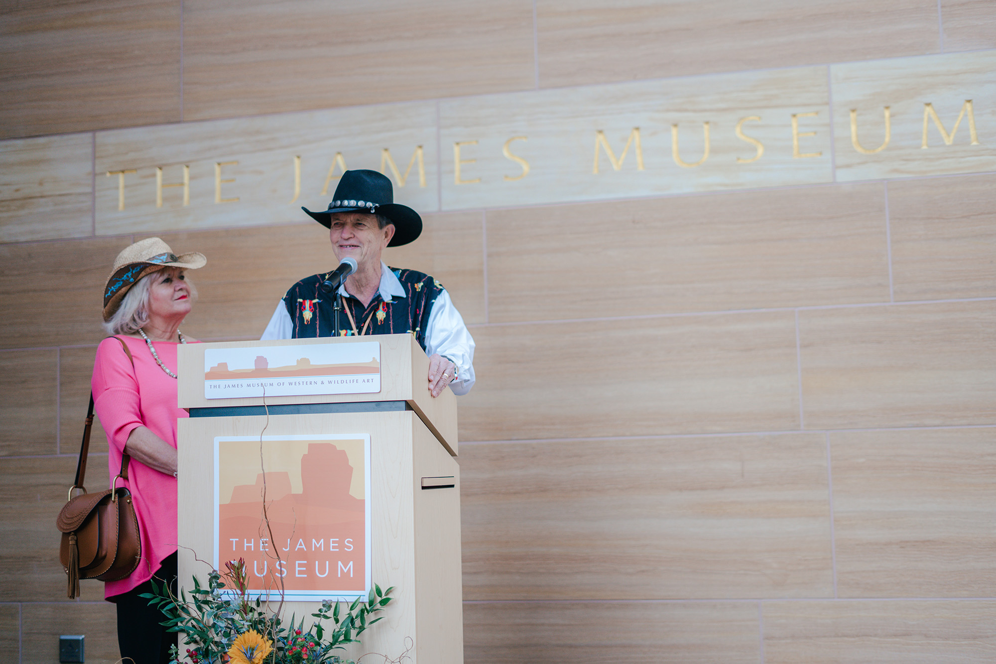 A man and woman in Western attire, possibly the museum's founders, stand at a podium with "The James Museum" sign, speaking in front of a wooden wall that highlights the institution's history.