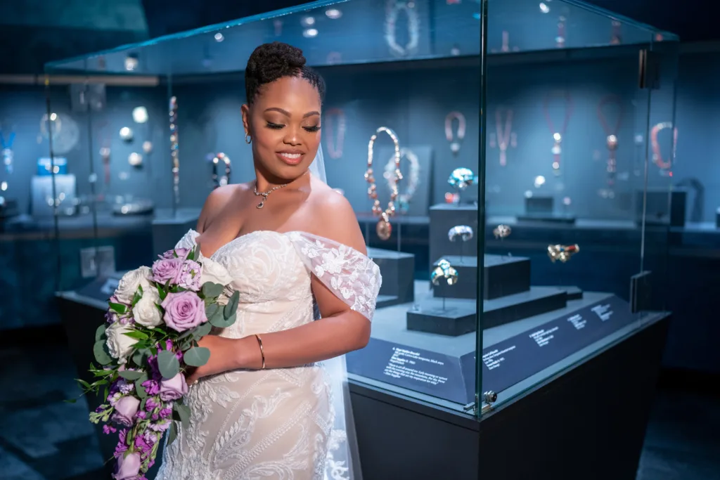 A bride in a white lace gown holds a bouquet of purple and white flowers, capturing the elegance of weddings as she stands near a glass jewelry display in a museum or gallery.