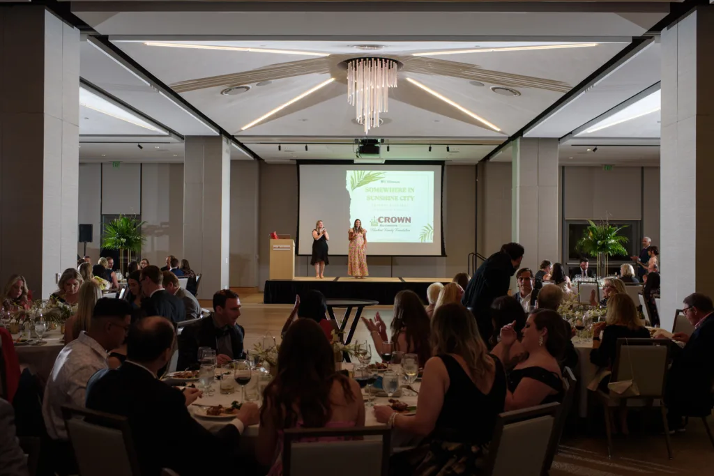 Guests seated at round tables attend a formal celebration in a banquet hall, while two speakers stand on stage in front of a large screen and podium—perfect for weddings or other grand occasions.