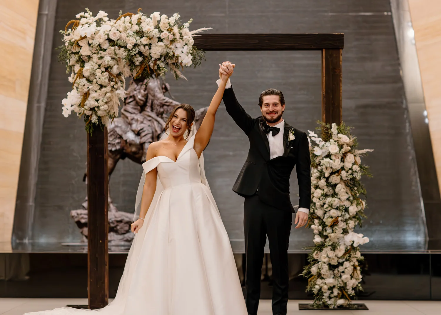 A bride and groom stand holding hands and smiling under a floral wedding arch indoors, capturing the joy of weddings and special celebrations.