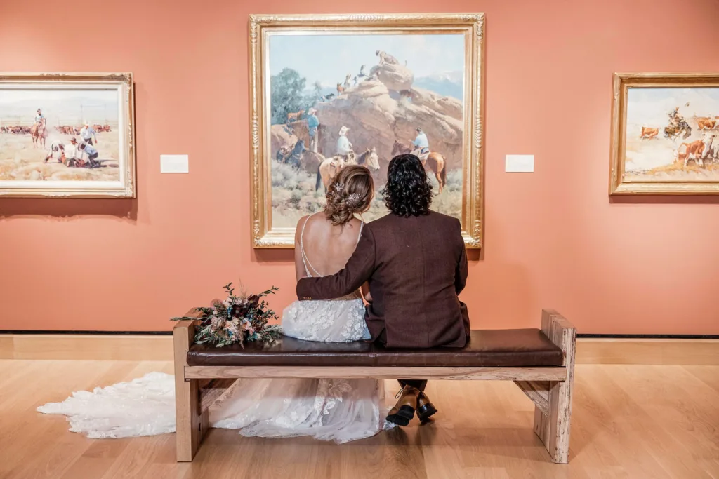 A bride and groom sit closely together on a bench, facing Western-themed paintings in an art gallery—capturing the quiet joy of weddings and intimate celebrations.