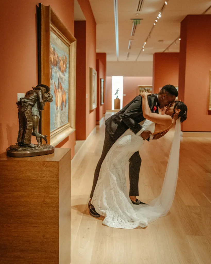A bride and groom share a kiss while the groom dips the bride in an art gallery with red walls, paintings, and a sculpture on display—capturing the elegance of weddings amid unique celebrations of art.