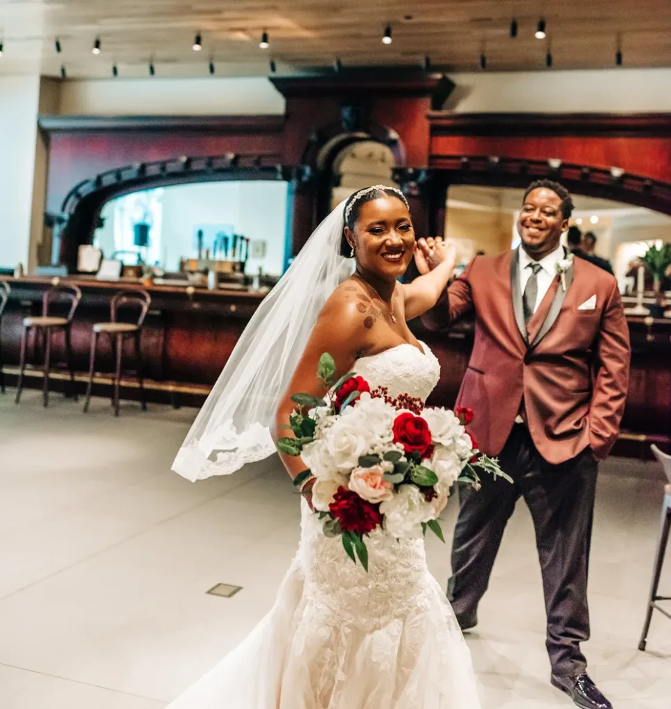 A bride in a white gown holding a bouquet and smiling, while a groom in a burgundy jacket stands behind her in a vibrant bar setting—capturing the joyful spirit of weddings and celebrations.