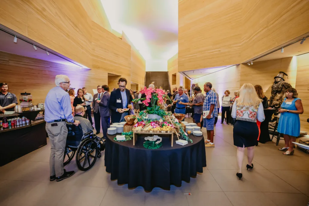 A group of people gather in a modern room around a round table decorated with flowers and food, engaging in lively conversation and enjoying refreshments during celebrations.