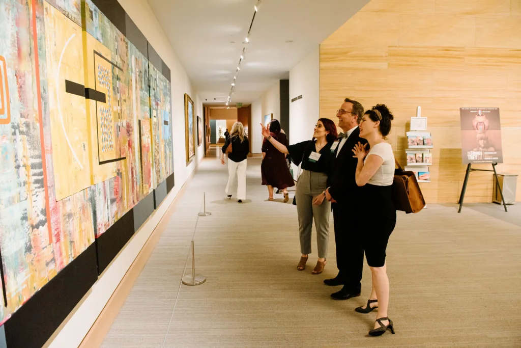 A group of people stand in a gallery hallway, observing and discussing a large abstract painting displayed on the wall as they reminisce about recent celebrations and weddings they've attended.
