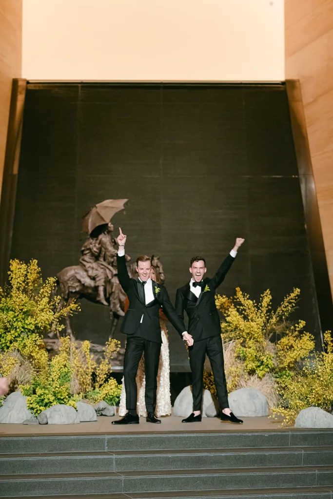 Two men in tuxedos stand on steps holding hands, both raising an arm in celebration, as they enjoy one of life's joyful weddings amid a large statue and lush greenery.