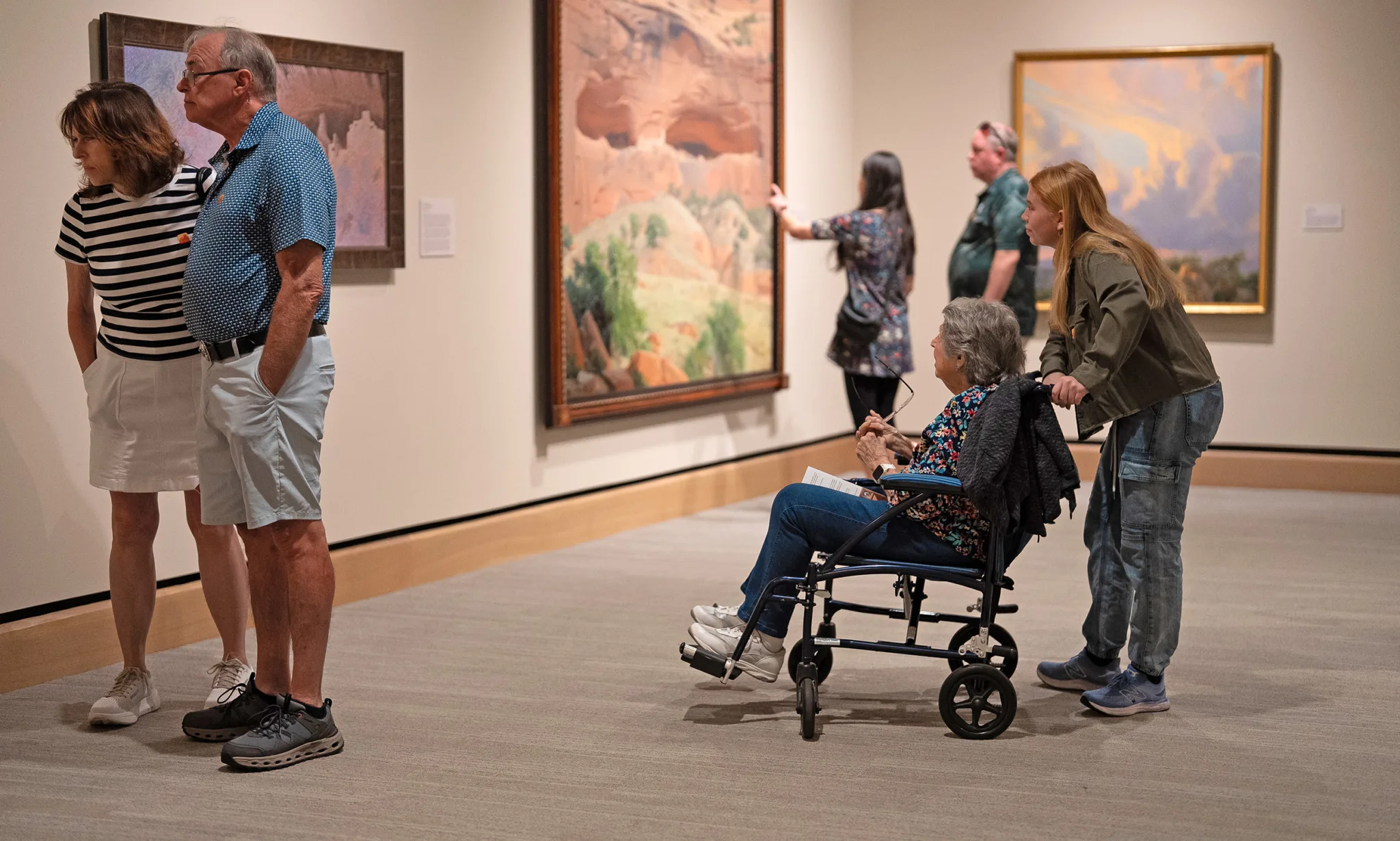 A group of people, including a woman in a wheelchair, view paintings on display in an art gallery featuring Accessibility Programs.
