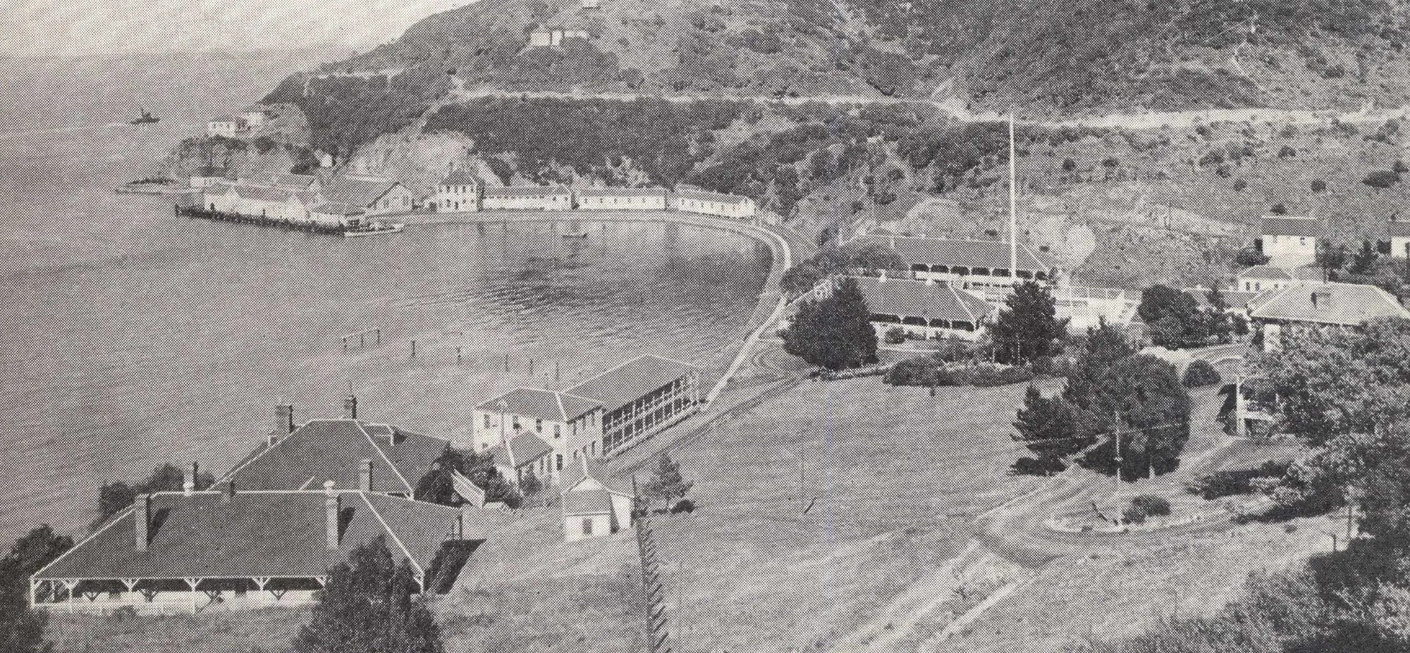 Black and white photo of a coastal settlement, featured in FarEast 08 Newspapers, with several buildings, a pier, and a hill rising in the background along the shoreline.