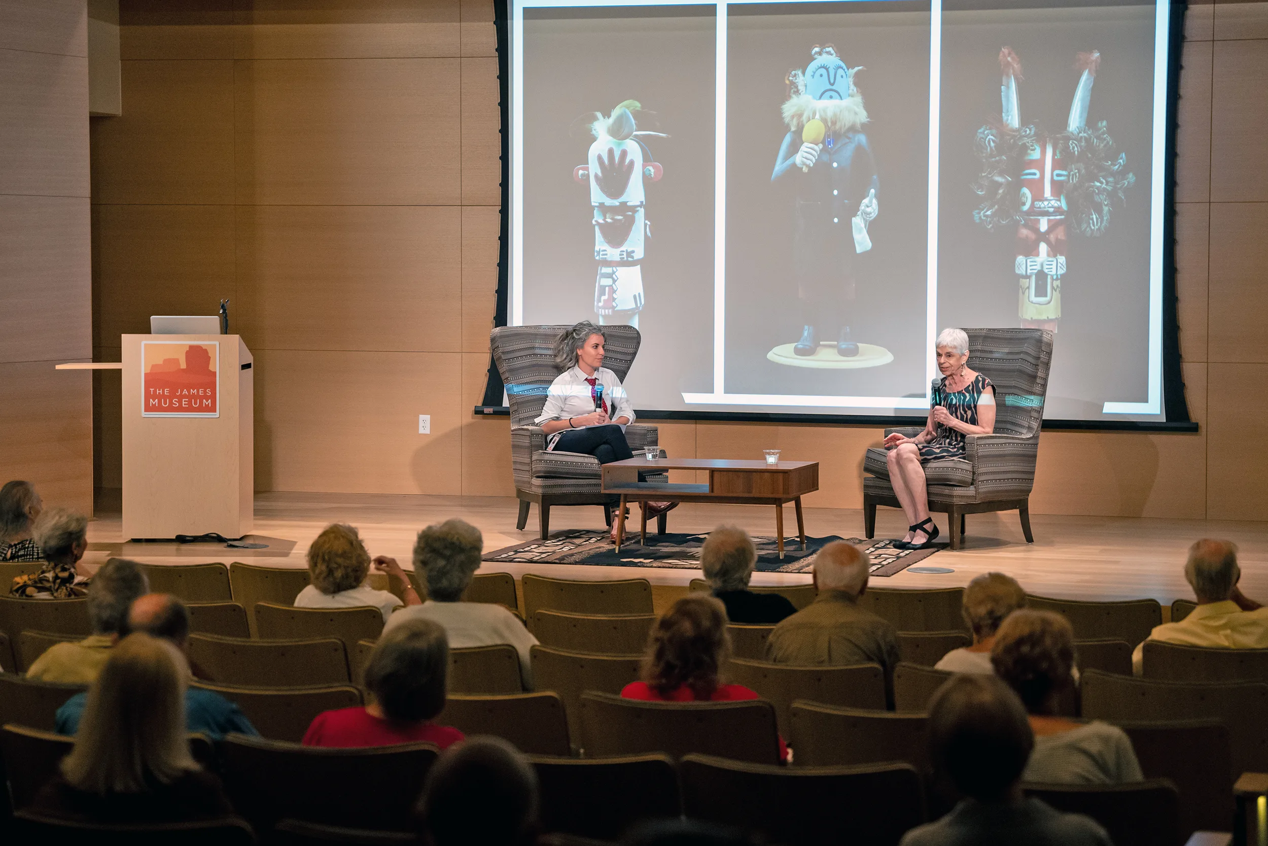 Two women converse on stage at The James Museum during an Adult Programs event, as an audience listens; three Native American figurines are displayed on a large screen behind them.