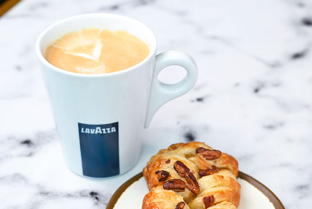A white Lavazza coffee mug filled with coffee sits on a marble surface next to a pastry with pecans on a small plate, highlighting the simple pleasures included in Member Benefits.