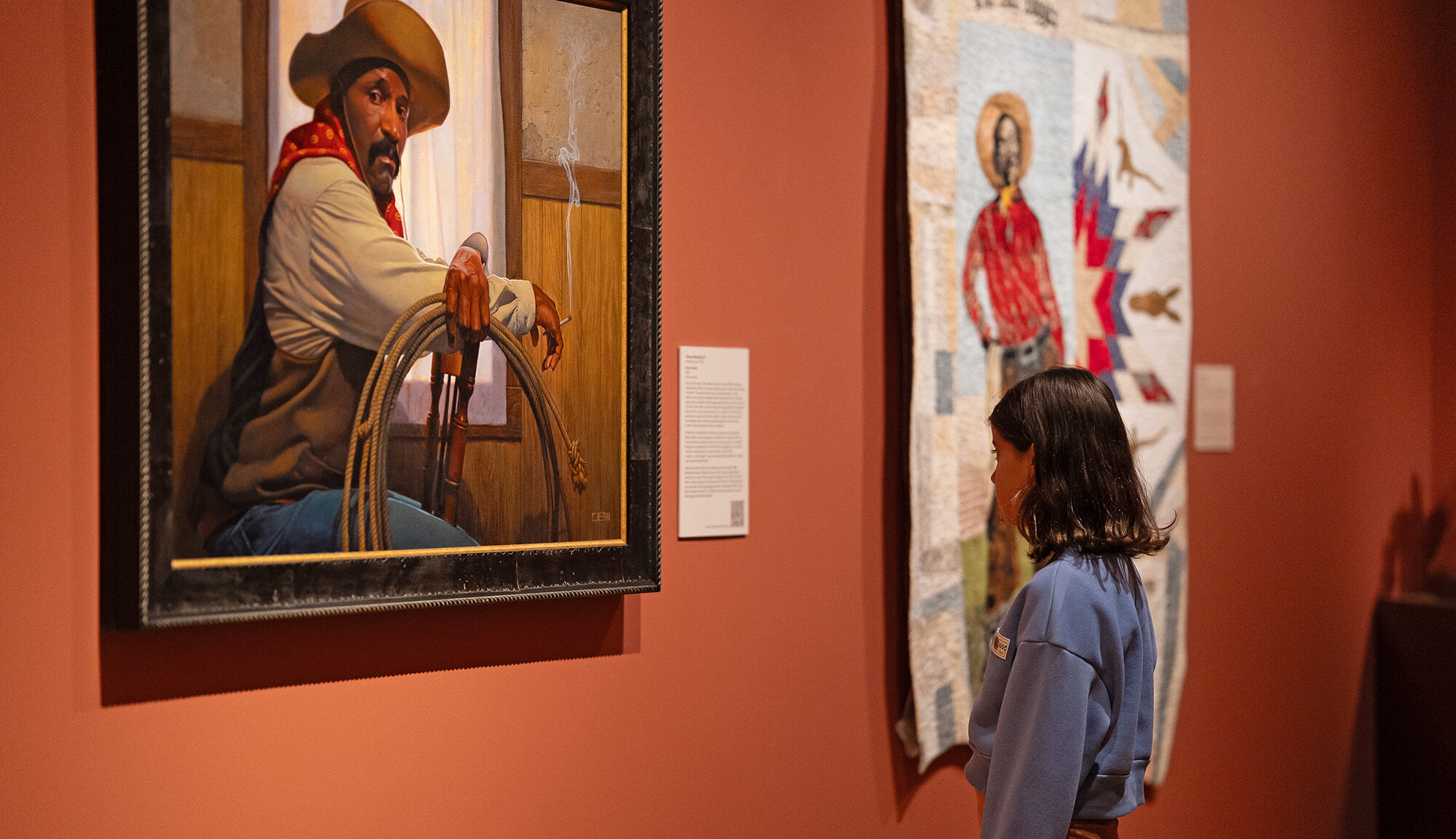 A person stands in a museum, contemplating the money value and artistry of a framed painting of a cowboy holding a lasso, with a textile artwork hanging on the adjacent wall.