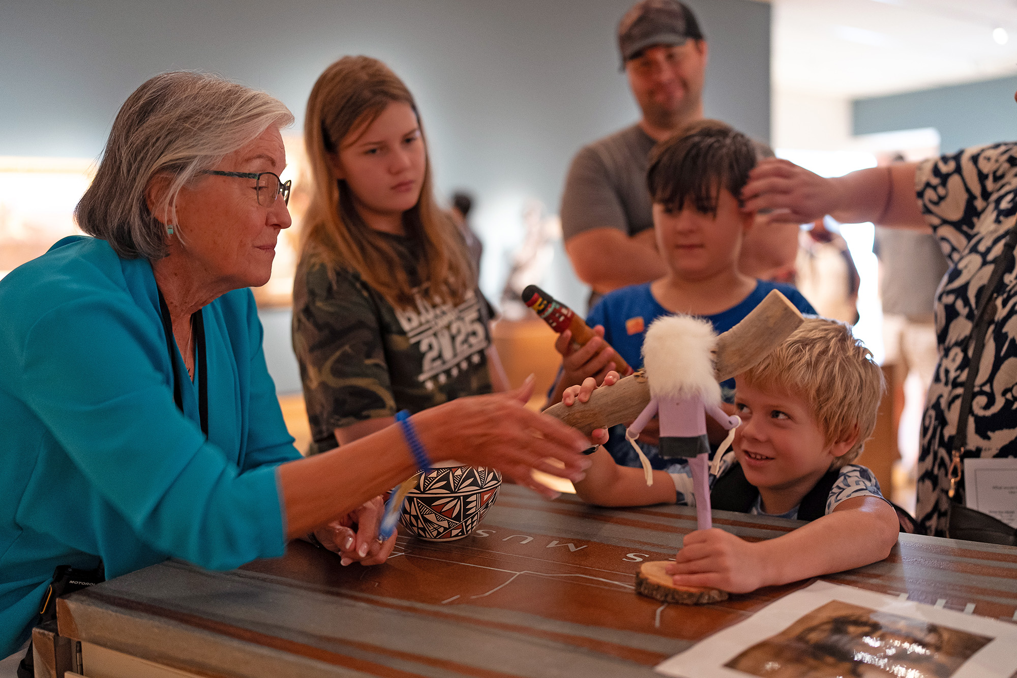 An older woman introduces a group of children and adults to various artifacts on a table in a museum, highlighting youth and family programs in an engaging setting.