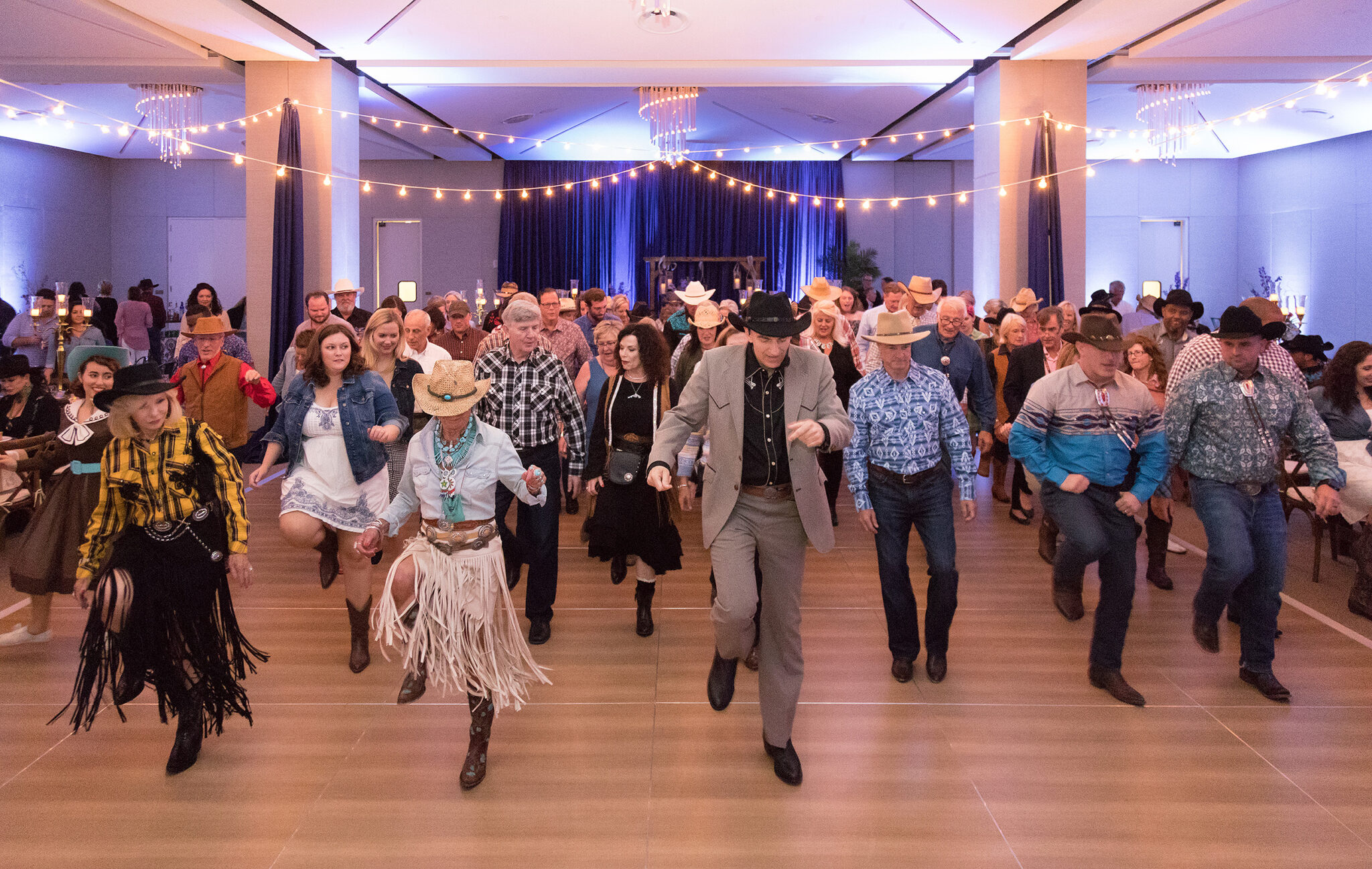 A group of people dressed in western attire line dancing at The James Jamboree in a large, well-lit indoor event space with string lights overhead.
