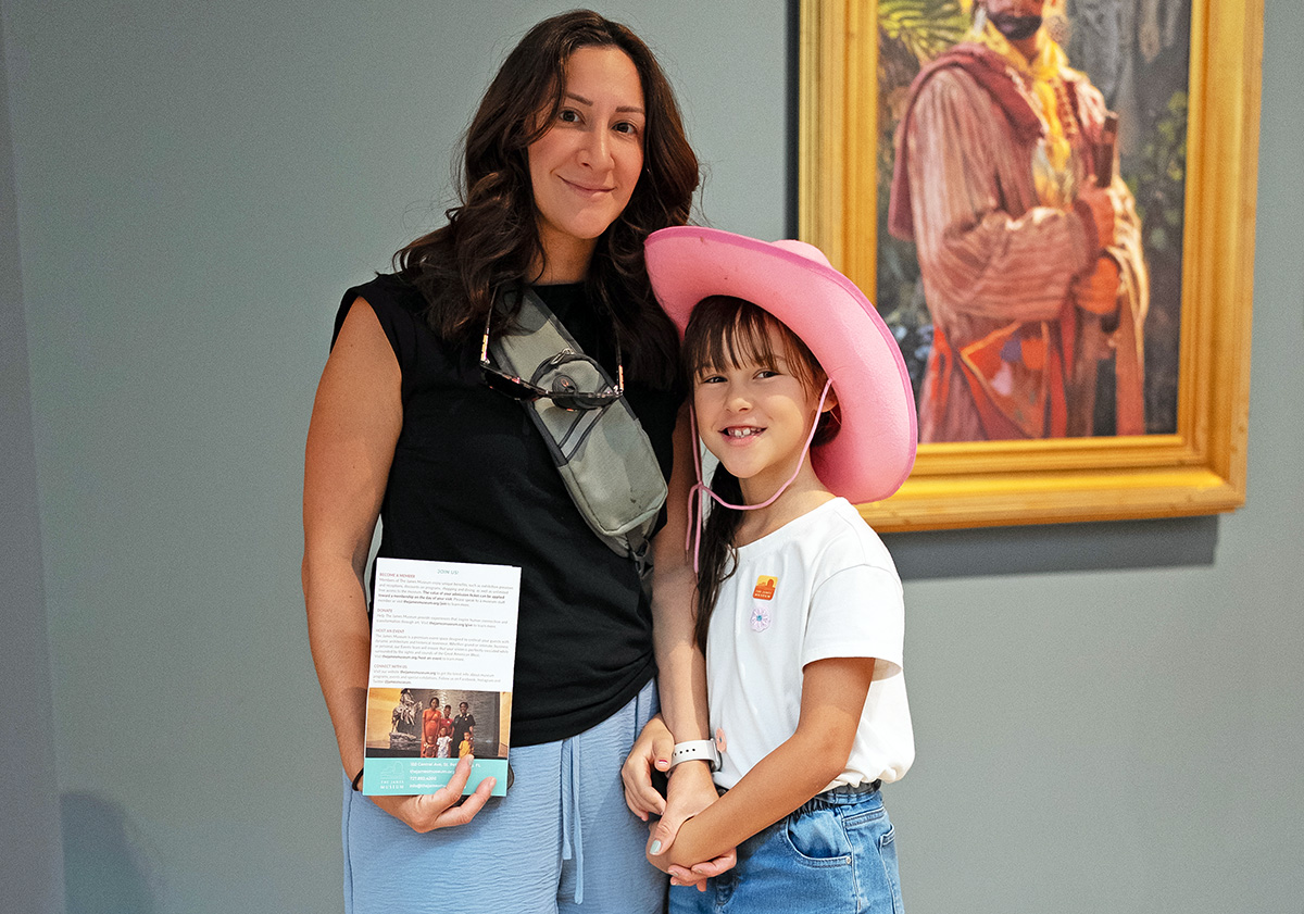 An adult and a child wearing a pink cowboy hat stand in front of a framed painting. The adult holds a brochure highlighting member benefits, and both are smiling at the camera.