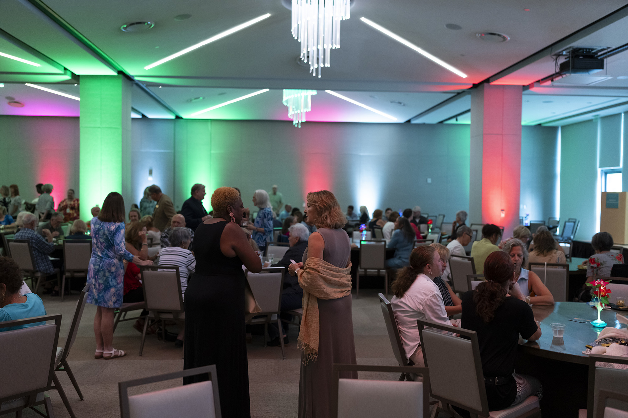 People are seated and standing in a large event room with colorful red and green lighting, a chandelier overhead, and a lively atmosphere perfect for memorable Member Events.