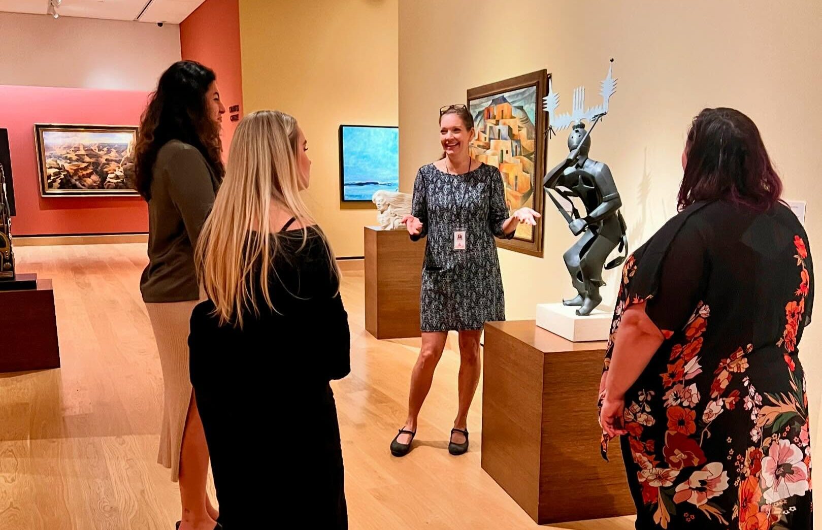 A museum guide speaks to three visitors in an art gallery, discussing the economic power of art while standing near paintings and a modern abstract sculpture.