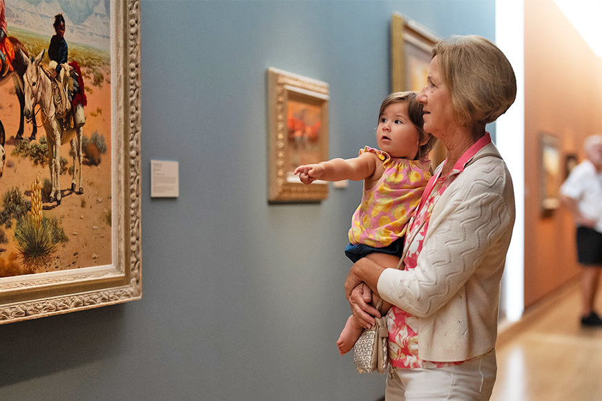 An older woman holds a young child who is pointing at a painting in an art gallery, highlighting the engaging family programs available. Other paintings and visitors are visible in the background.