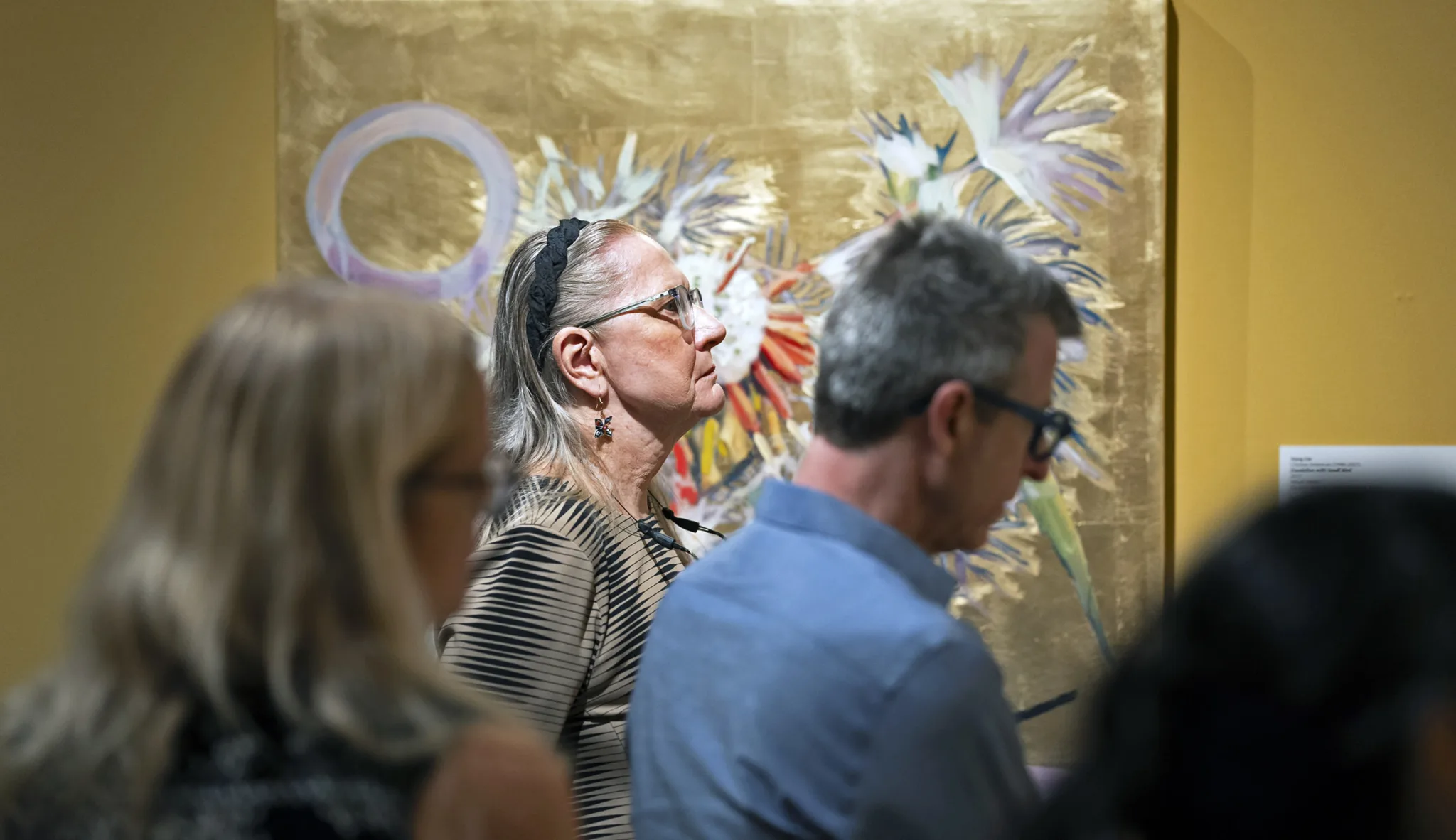 A group of people stand and observe an abstract painting with gold and colorful floral elements, part of the FarEast 12 collection, as they contemplate its reflections in an art gallery.